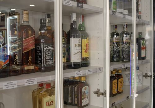 Estante de bebidas alcohólicas con varias botellas de ron, whisky, vodka y licor, exhibidas en un mueble con puertas de vidrio. Los precios están claramente etiquetados en cada estante.