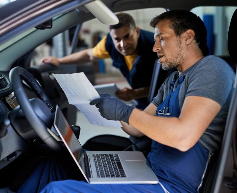 Técnico automotriz revisando documentos y utilizando una computadora portátil dentro de un vehículo, mientras otro colega observa en un taller de reparación.